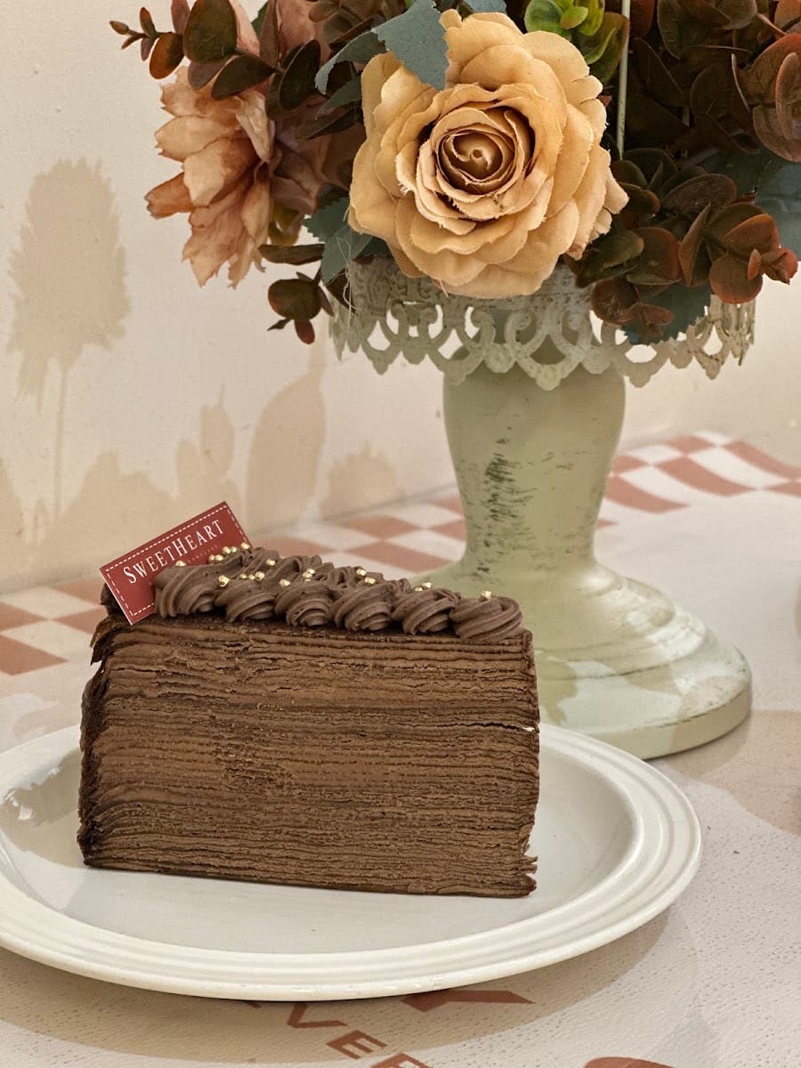 A slice of layered chocolate cake with a Sweetheart tag, in front of a vase of roses