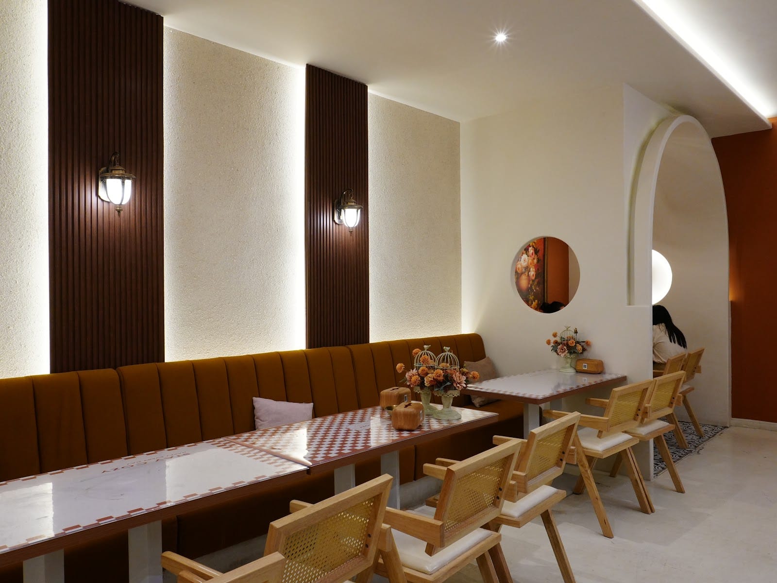 Inside Sweetheart Cafe: a long banquette, rattan chairs and checkered tables under wall lamps, with an arched doorway and a round window